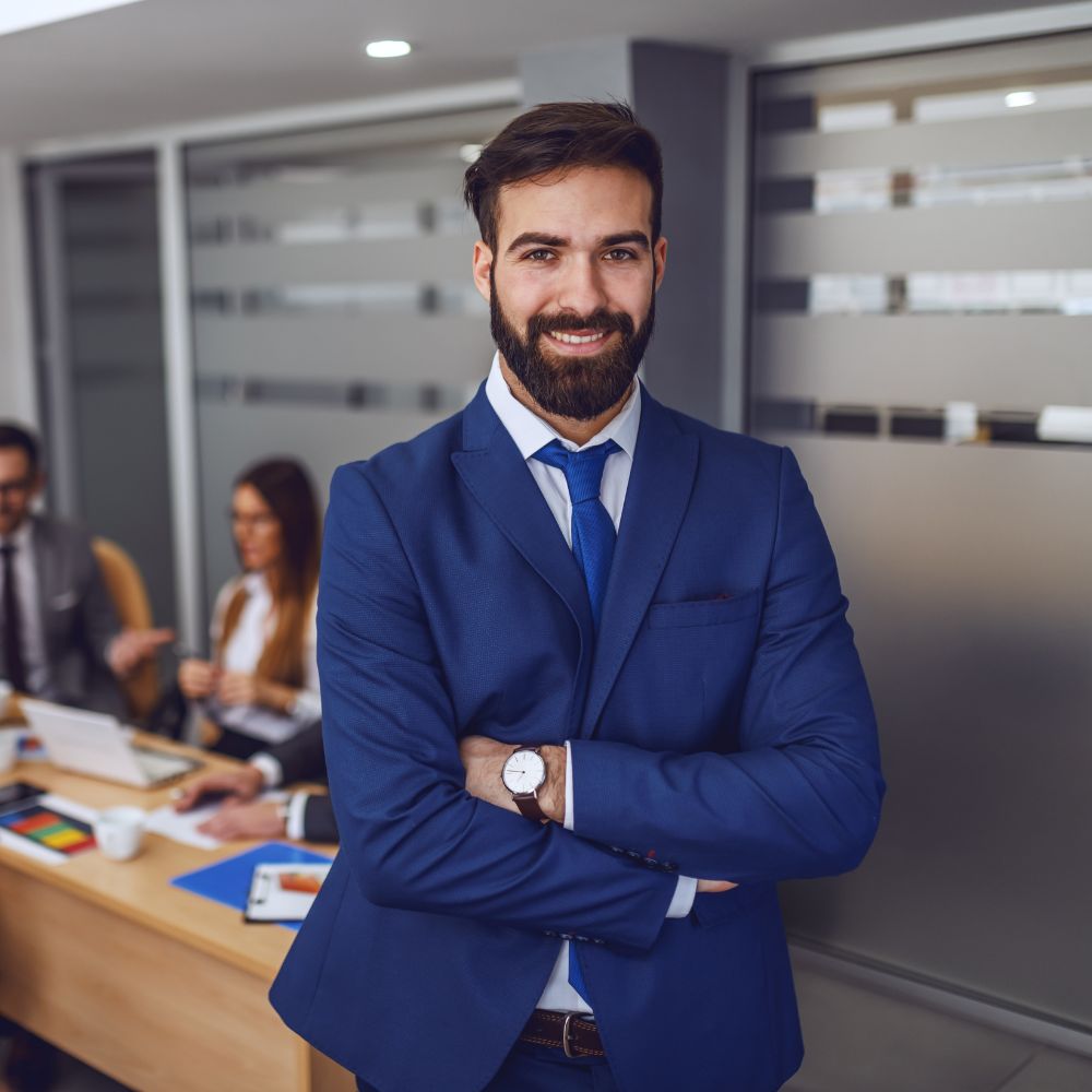 Professionele man in blauw pak glimlachend vol vertrouwen in een kantooromgeving met collega's op de achtergrond.