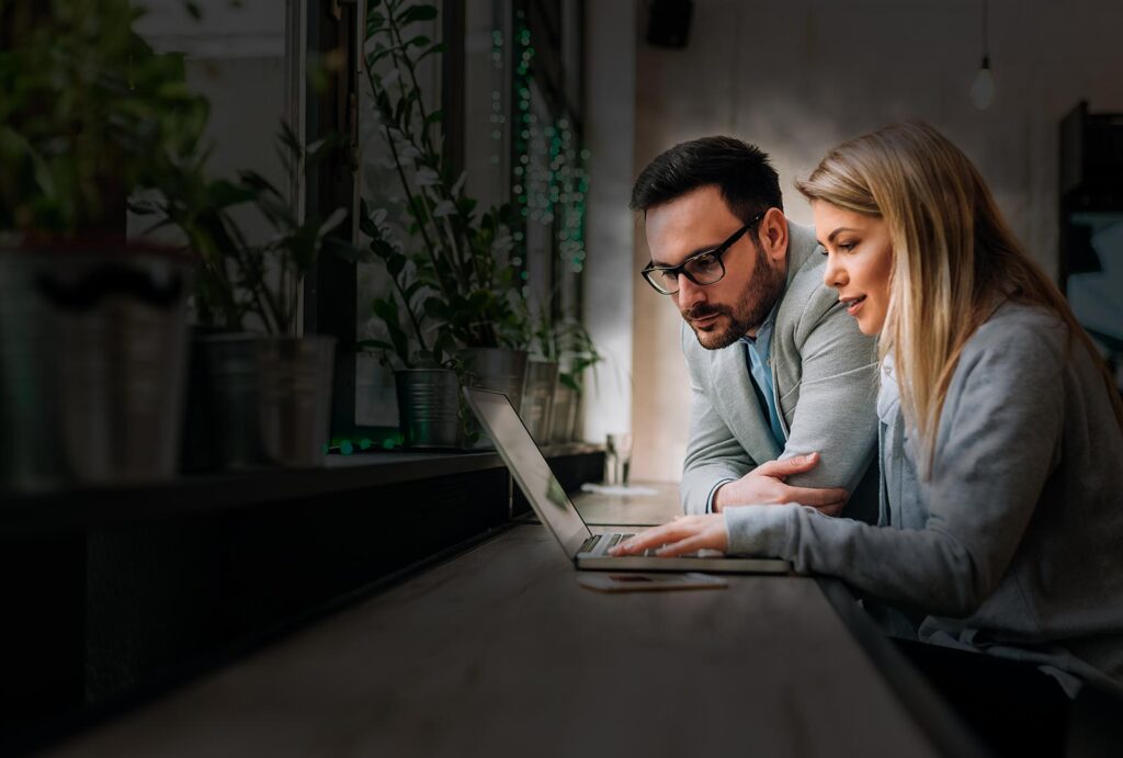 Twee professionals, een man en een vrouw, bekijken aandachtig de inhoud op een laptop in een moderne kantooromgeving met planten.