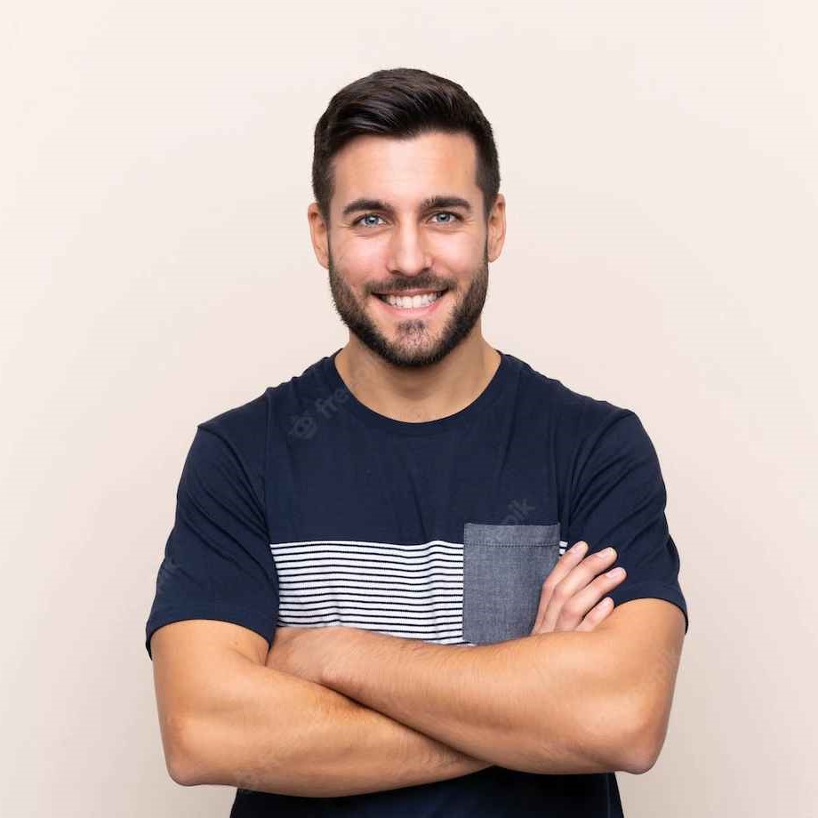 Een jonge man met een baard, glimlachend, gekleed in een blauw gestreept T-shirt, armen gekruist, staande tegen een lichtbeige achtergrond.