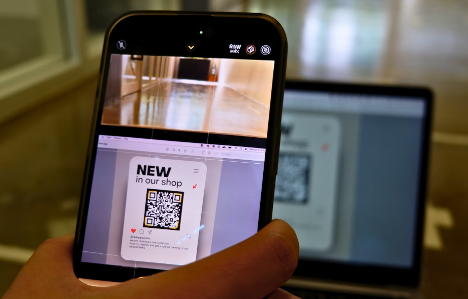 De hand van een persoon houdt een smartphone vast en scant een QR-code op het scherm van een laptop om nieuwe artikelen in een winkel te promoten met behulp van quishing-technieken.