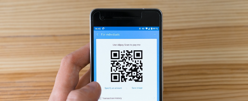 Hand die een smartphone vasthoudt met een QR-code voor een quishing-aanval op het scherm, tegen de achtergrond van een houten tafel.