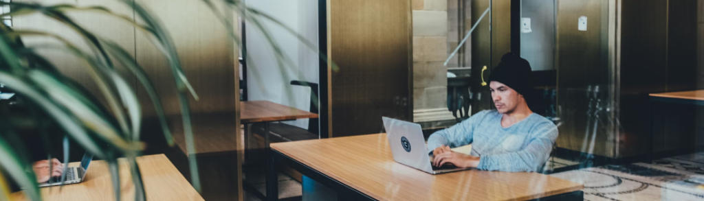 Een jonge man met een muts en een blauwe trui geconcentreerd aan het werk op een laptop aan een houten tafel in een moderne kantoorruimte gericht op bedrijfsbeveiliging.