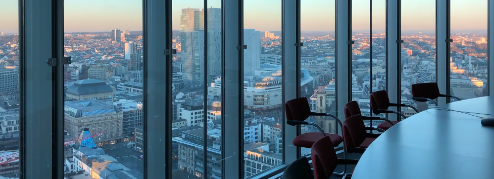 Een stadsgezicht gezien vanuit de beveiligde vergaderruimte van een hoog gebouw, met een lange tafel met stoelen en grote ramen die gebouwen tegen een zonsondergang laten zien.