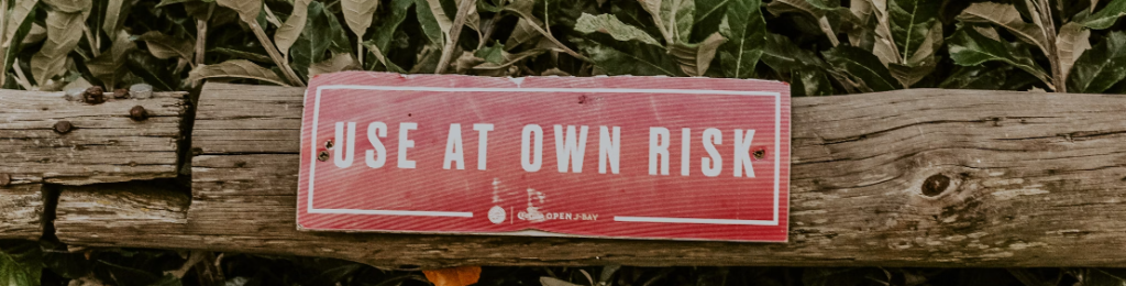 Een vervaagd rood bord 'Gebruik op eigen risico' bevestigd aan een verweerde houten balk met een groene bladachtergrond, een voorbeeld van risicomanagement in zijn omgeving.