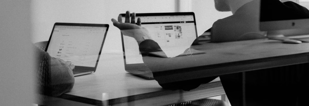 Een man werkt aan een bureau met drie computerschermen en concentreert zich met zichtbare concentratie op taken die verband houden met cybersecurity. De foto is in zwart-wit.