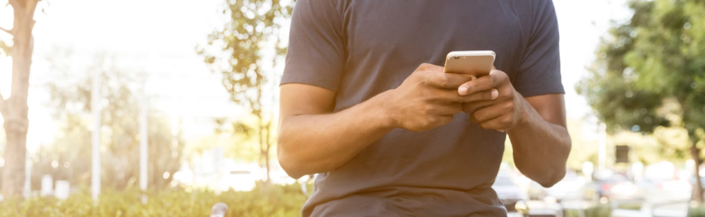 Man in een blauw t-shirt die buitenshuis een smartphone gebruikt terwijl zonlicht door de bomen filtert, op zijn hoede voor mogelijke phishing-aanvallen.