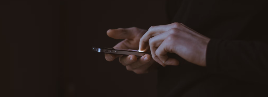 Handen van een persoon in donkere kleding die een smartphone gebruikt in een slecht verlichte omgeving om meer te weten te komen over vormen van phishing.