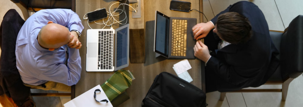 Twee mensen werken aan een tafel met laptops, telefoons en papieren. De één is kaal en de ander heeft donker haar. Ze zitten tegenover elkaar, beiden gefocust op hun taken, en zorgen er wellicht voor dat hun systemen beveiligd zijn tegen phishing-pogingen terwijl ze ijverig werken.