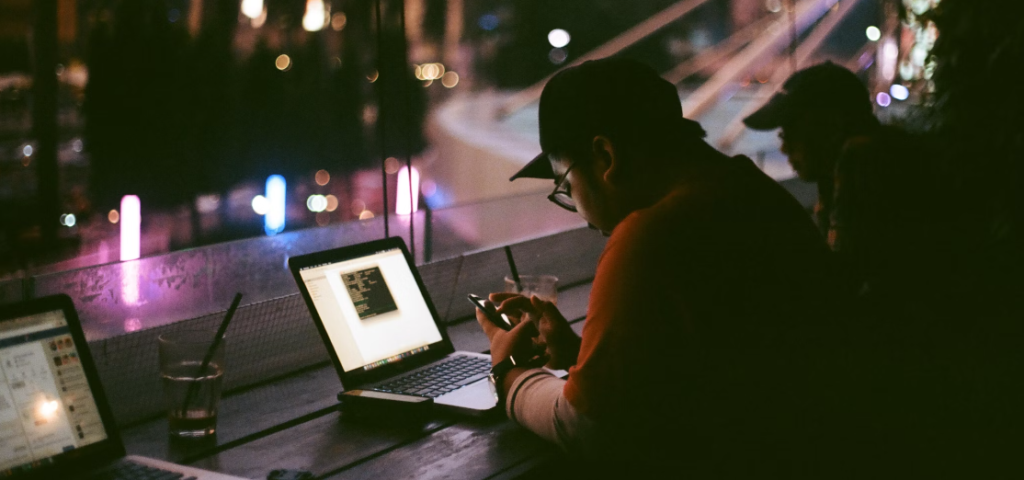 Een persoon zit 's nachts aan een bar, werkt op een laptop met een smartphone in de hand, omringd door straatverlichting en alert op vormen van phishing-bedreigingen.