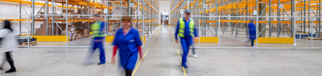Werknemers in veiligheidsvesten lopen door een groot magazijn met hoge oranje planken, waarvan één een schoonmaakmop voortduwt.