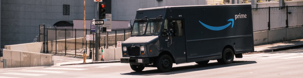 Een Amazon Prime-bestelwagen rijdt door een stedelijk gebied met verkeerslichten en gebouwen op de achtergrond.