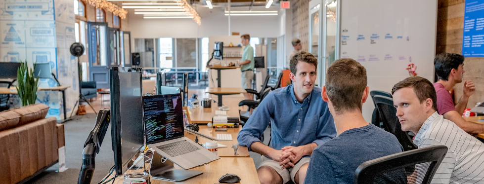 Mannen bespreken tegenstandersimulatie in een druk open kantoor met bureaus, computers en een informele zithoek.