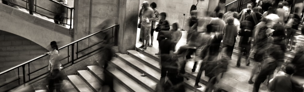 Een zwart-witfoto van een druk binnentrappenhuis waar veel mensen de trap op en af lopen, waarvan sommige wazig bewegen, wat doet denken aan de constante drukte en waakzaamheid in een Security Operations Center.
