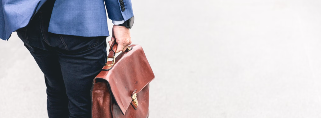 Man in een blauwe blazer met een bruinleren aktetas op een lichtgrijze achtergrond in het Security Operations Center (SOC).