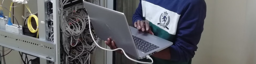 Persoon in een blauw sweatshirt met een laptop naast een serverrek en rommelige kabels in een netwerkruimte, die de Top 10 Cybersecurity Trends voor 2024 analyseert.