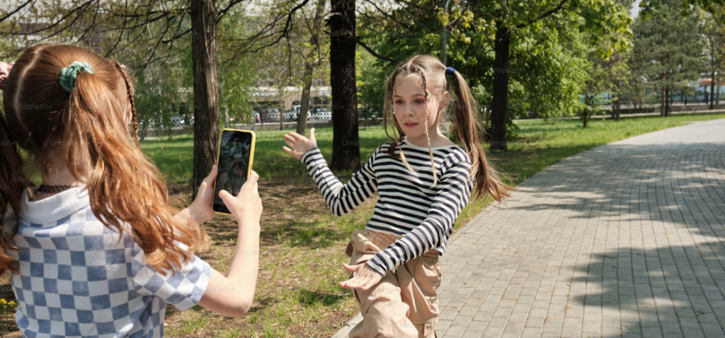 Een meisje maakt met een smartphone een foto van haar vriendin die langs een parkpad danst. Heldere daginstelling. De afbeelding kan onbedoeld bijdragen aan cyberpesten als deze op onverantwoorde wijze online wordt gedeeld.
