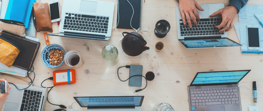 Bovenaanzicht van een drukke werkruimte met meerdere mensen die op laptops typen, omringd door verschillende digitale apparaten, beveiligde poort, notebooks en kopjes.