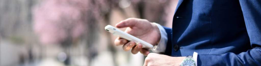 Een persoon in een blauw pak houdt buiten een witte smartphone vast, met een onscherpe achtergrond van roze bomen. In een tijdperk waarin telefoons wijdverspreid zijn, blijven de zorgen over spyware onder veel politici heersen.