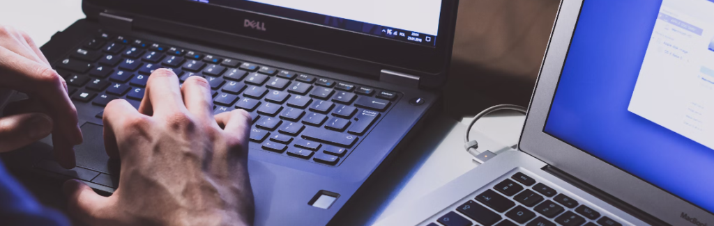 Twee laptops op een bureau. Iemand typt op het toetsenbord van de ene laptop, gefocust op een cybersecuritytaak, terwijl het scherm van de andere laptop een wazige interface weergeeft, wat bijdraagt aan een efficiënt digitaal ecosysteem.