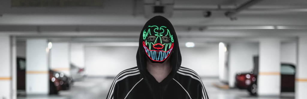 Persoon met een zwarte hoodie en een kleurrijk, verlicht masker in een parkeergarage, die als bewaker van cyberveiligheid naadloos opgaat in het stedelijke ecosysteem.