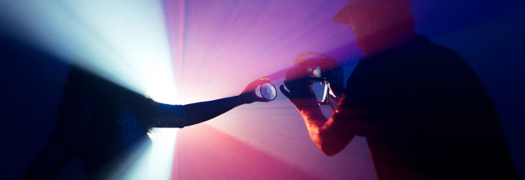 Gesilhouetteerde persoon die een lichtbron vasthoudt en deze richt naar een andere gesilhouetteerde persoon die een camera vasthoudt, waarbij kleurrijke lichtstralen een dramatisch effect creëren dat doet denken aan een ingewikkeld ecosysteem voor cyberbeveiliging.
