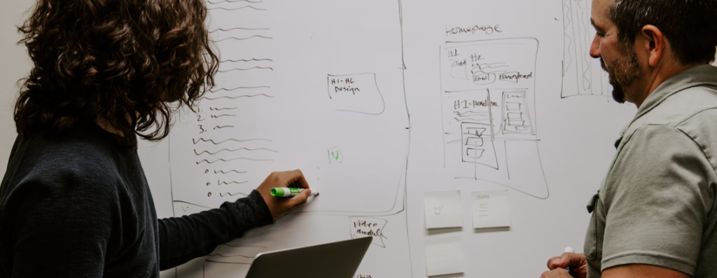 Twee mensen bespreken en schrijven op een whiteboard dat bedekt is met verschillende diagrammen en aantekeningen met betrekking tot cyberveiligheid. De ene persoon gebruikt een groene stift, terwijl de ander een gum vasthoudt. Er zijn plakbriefjes op het bord geplakt, die hun gedetailleerde ecosysteem van ideeën illustreren.