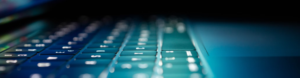 Close-up zijaanzicht van een verlicht, slanker toetsenbord met blauwe en groene achtergrondverlichting; de toetsen zien er scherp uit tegen een donkere achtergrond en stralen een strak ontwerp uit dat populair is onder hackers.