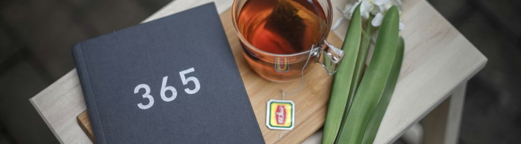 Een strak zwart notitieboekje met "365" op de omslag, een glazen kopje thee, een theezakje en twee witte bloemen op een houten dienblad.
