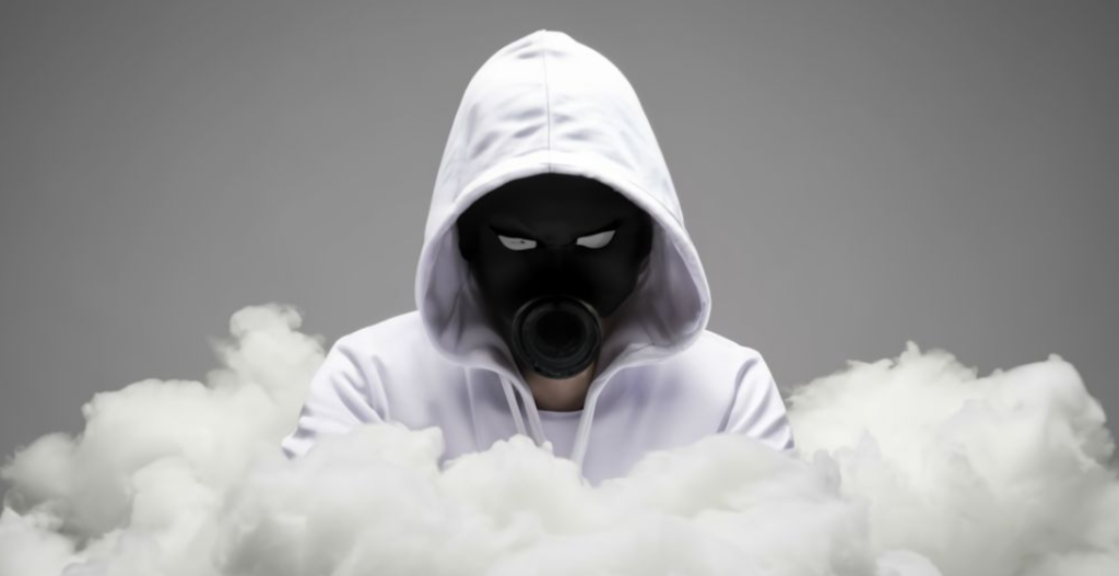 Een persoon met een witte hoodie en een zwart gasmasker, die doet denken aan anonieme hackers, staat omringd door rook tegen een grijze achtergrond.