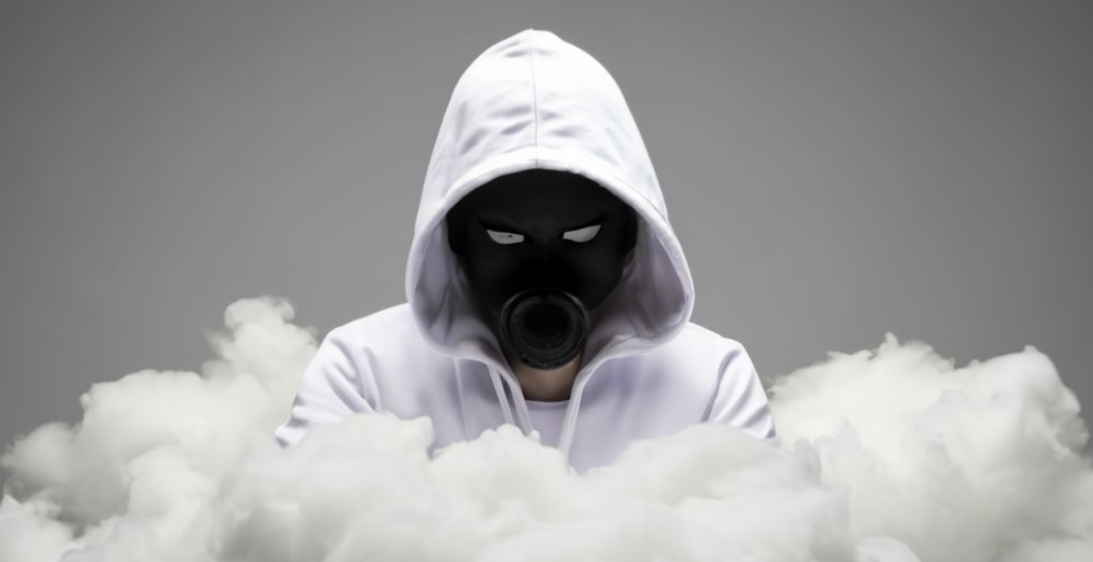 Een persoon met een witte hoodie en een zwart gasmasker, die doet denken aan anonieme hackers, staat omringd door rook tegen een grijze achtergrond.