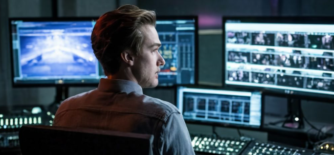 Man houdt toezicht op meerdere bewakingsschermen in een donkere controlekamer gericht op cyberveiligheid.