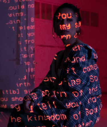 Een vrouw in een geruit pak zit onder rood licht en projecteert met grote impact tekst op haar gezicht en lichaam.