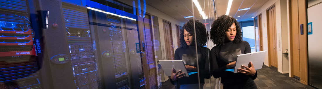 Een vrouw met een tablet inspecteert serverracks in een datacenter en weerspiegelt de Cyberveiligheidsmaatregelen op het glaspaneel voor haar.