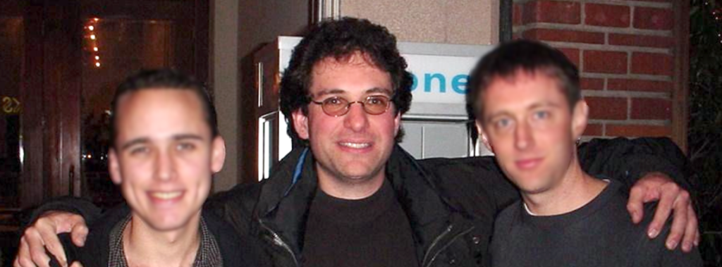 Onder de sterrenhemel poseren drie mensen voor een foto buiten een gebouw. Ze spelen op subtiele wijze in op de mysterieuze uitstraling van de beroemde hacker Kevin Mitnick en zijn wereld van cybersecurityintriges.