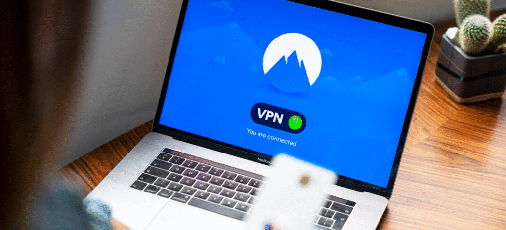 Een laptop toont een vpn verbindingsscherm met de boodschap "u bent verbonden", terwijl een persoon een smartphone voor zich houdt op een houten bureau, waarmee het belang van beveiliging wordt benadrukt bij het gebruik van openbare wifi netwerken, die gevaarlijk kunnen zijn.