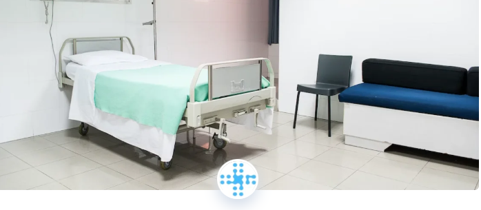 Een ziekenhuiskamer met een eenpersoonsbed, witte lakens en een mintgroene deken, naast een stoel en een blauwzwarte bank tegen een witte muur.