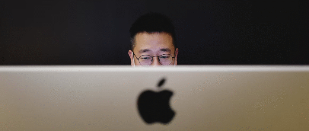 Een persoon met een bril is gedeeltelijk zichtbaar achter een apple computer, wat duidt op een bezorgdheid over icloud beveiliging, met een donkere achtergrond die het gevoel van privacy en veiligheid versterkt.