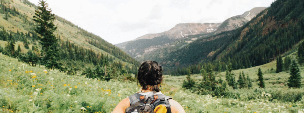 Persoon met rugzak staat in een groene vallei omringd door pijnbomen en bergen onder een bewolkte hemel.